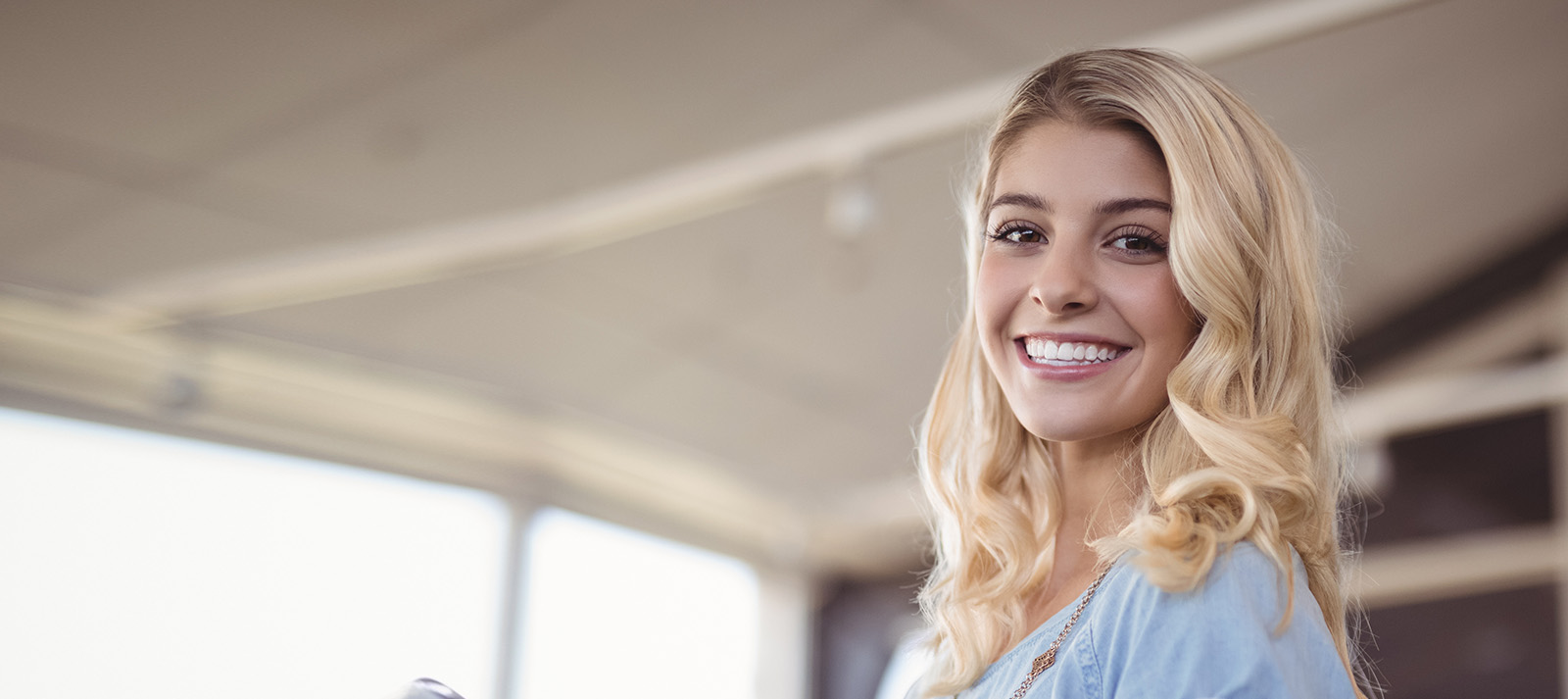 The image shows a smiling woman with blonde hair, wearing a light blue top, standing in an interior space that appears to be an office or lobby, looking directly at the camera.