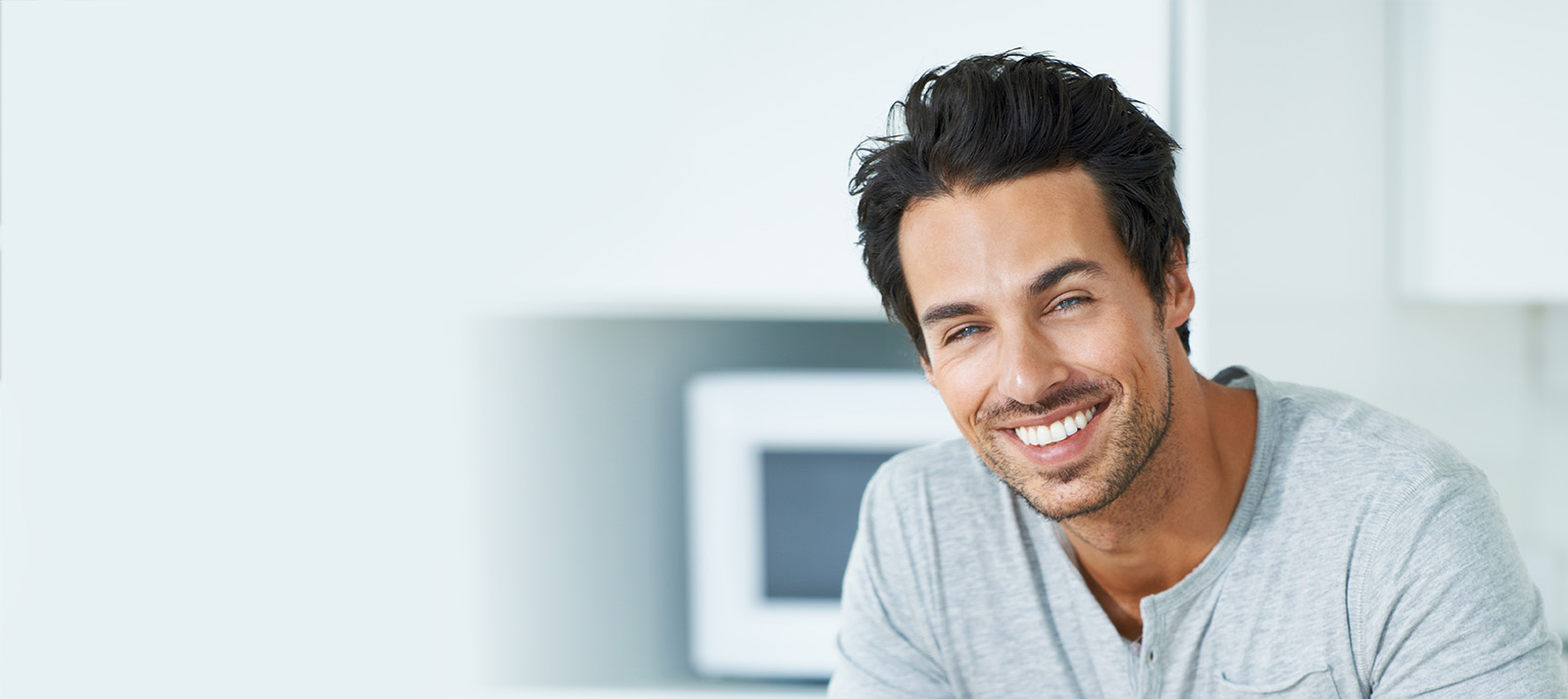 The image is a photograph of a man with short hair, wearing a light grey shirt, smiling at the camera. He appears to be in a kitchen setting, with a blurred background that suggests domestic appliances and countertops.