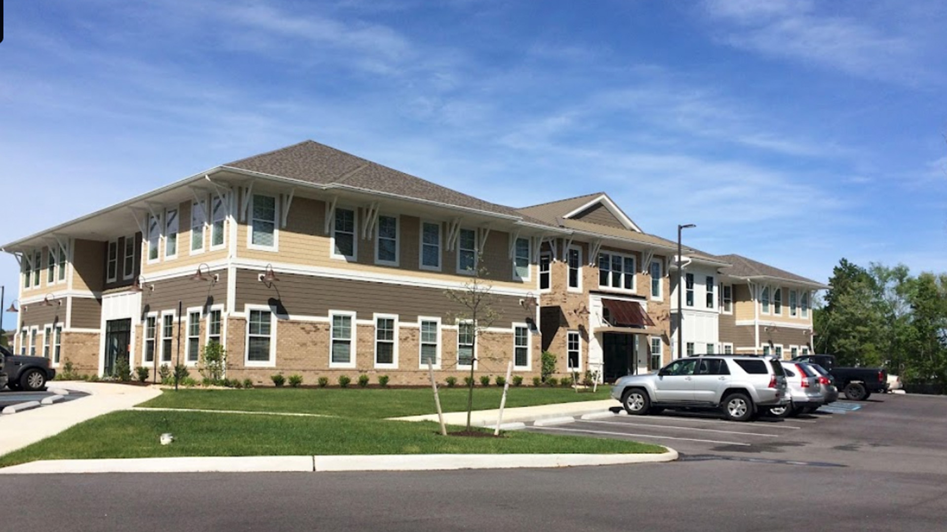The image shows a two-story building with a beige exterior and red trim, featuring multiple windows, a front door, and a covered porch. There is a parking lot in the foreground with several cars parked. The sky is clear, suggesting it might be midday or early afternoon.