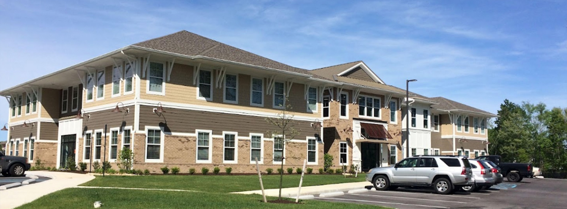 The image shows a two-story building with a beige exterior, featuring a flat roof and a front entrance with a covered walkway. The architecture suggests a modern residential or small commercial structure.