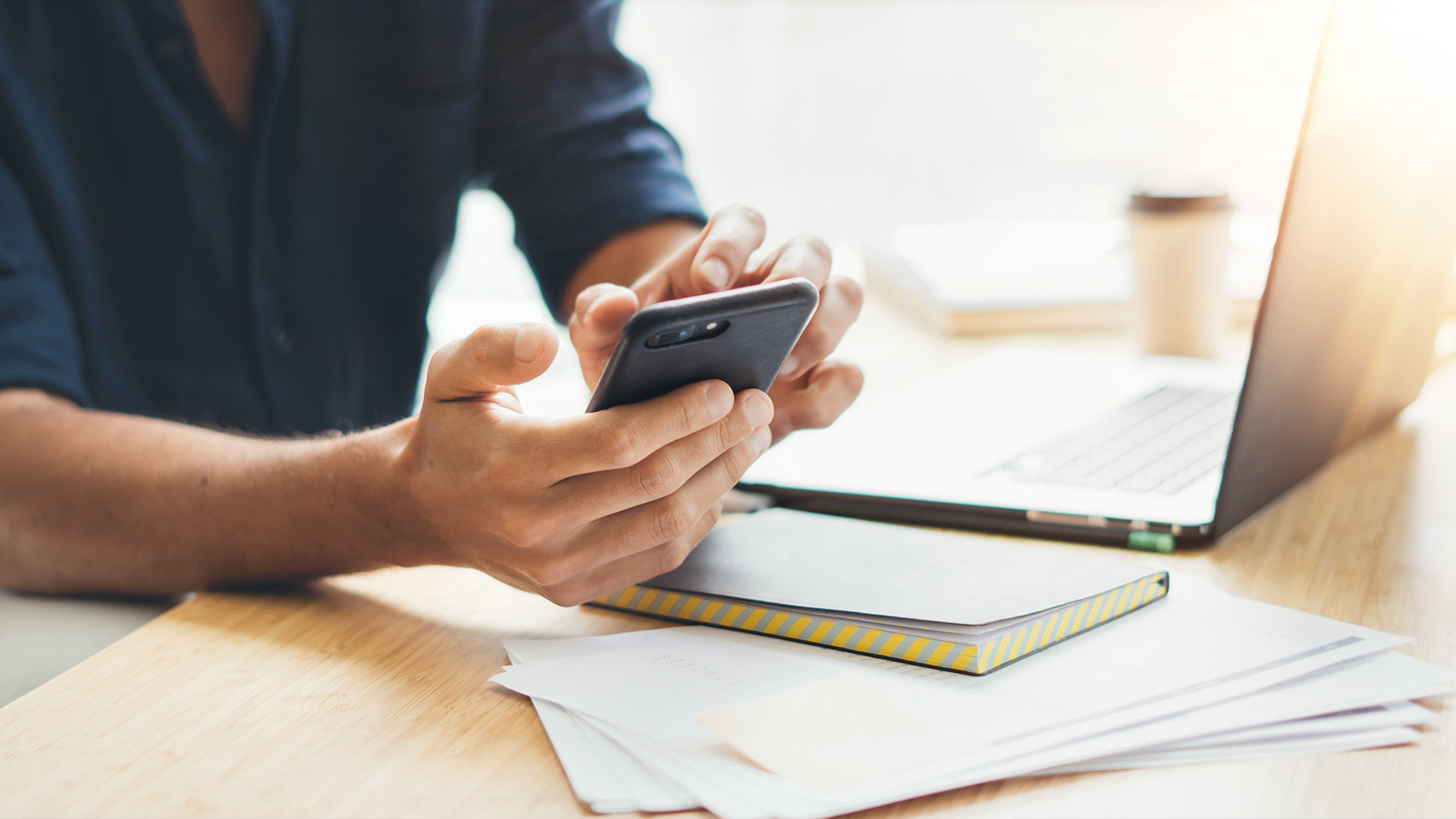 The image shows a person using a smartphone, with the device in their hands, while sitting at a table with various items such as notebooks and a laptop.