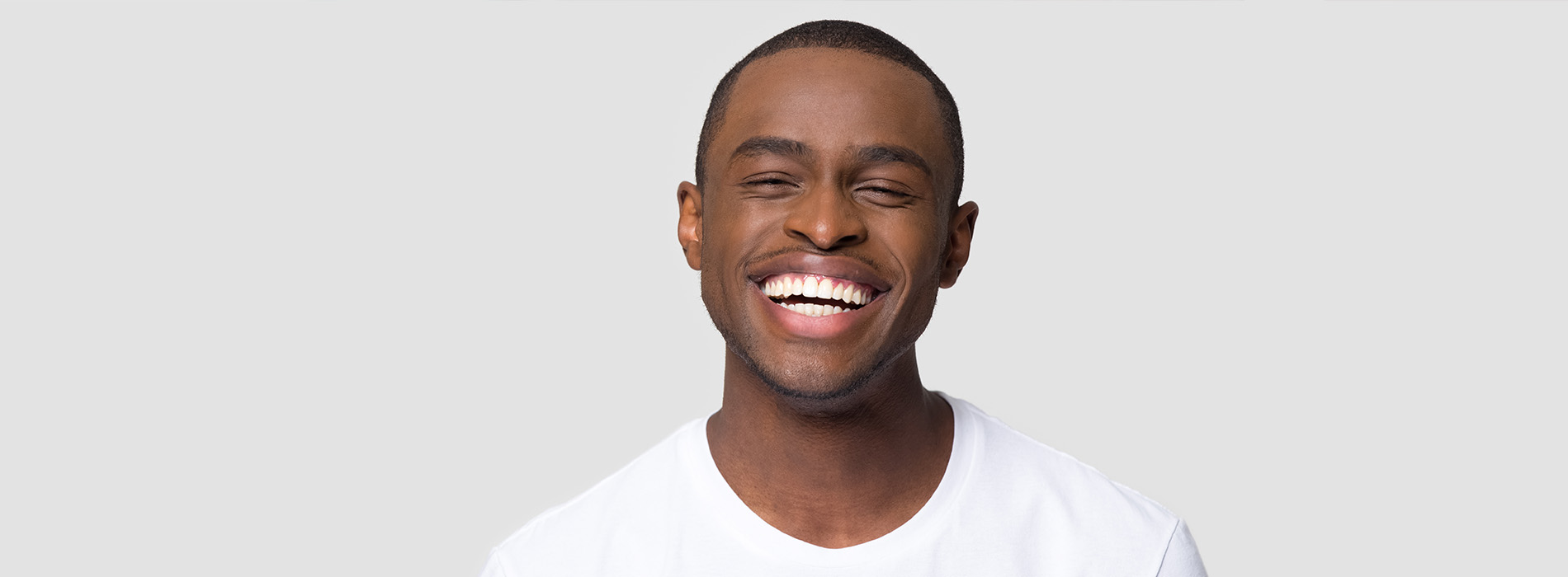 The image is a photograph of a smiling man with dark skin, wearing a white t-shirt. He appears to be in his twenties or thirties and has short hair. The background is neutral, emphasizing the subject.