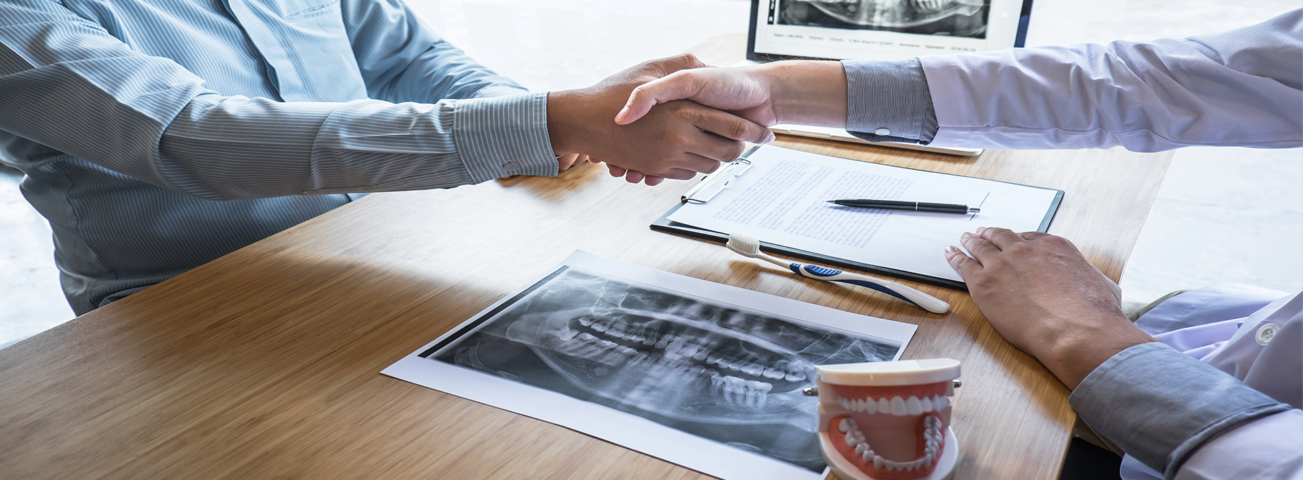 Two individuals shaking hands over a table with a laptop and papers, in an office setting.