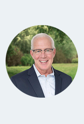 The image shows a man wearing glasses and a suit, smiling at the camera. He has white hair and appears to be middle-aged or older.