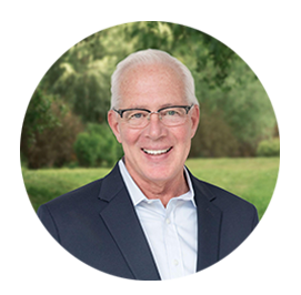 The image is a portrait of an older man with glasses, smiling at the camera. He has gray hair and is wearing a dark suit jacket over a light shirt. The background is out of focus but appears to be an outdoor setting with greenery.